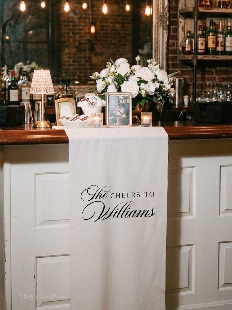 May include: A wedding bar is decorated with a white table runner bearing the words "The Cheers to Williams". The bar is decorated with white flowers, framed photos, candles, and a small lamp. The background features a brick wall.