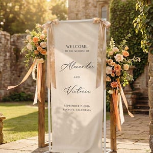 Könnte beinhalten: Ein vertikales, cremefarbenes Stoff-Willkommensschild für Hochzeiten in einem weißen Metallrahmen. Der Text lautet: WELCOME TO THE WEDDING OF Alexander AND Victoria SEPTEMBER 7, 2026 SANTA FE, CALIFORNIA. Zwei beige Bänder und Blumen schmücken den oberen Teil.