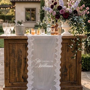 May include: A wooden bar with a white lace runner bearing the text "The Cheers to Williams." The bar is set up outdoors and features cocktails, bottles of spirits, champagne flutes, and a floral arrangement.