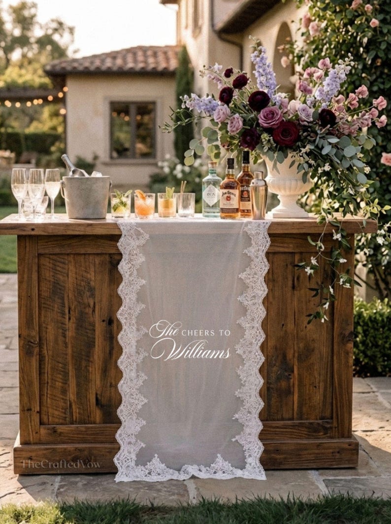 May include: A wooden bar with a white lace runner bearing the text "The Cheers to Williams." The bar is set up outdoors and features cocktails, bottles of spirits, champagne flutes, and a floral arrangement.