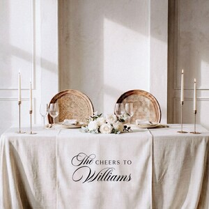 May include: A wedding table setting with a cream tablecloth. Two rattan chairs are placed opposite each other. The table is decorated with white roses, candles, and wine glasses. The text "The Cheers to Williams" is printed on the tablecloth.
