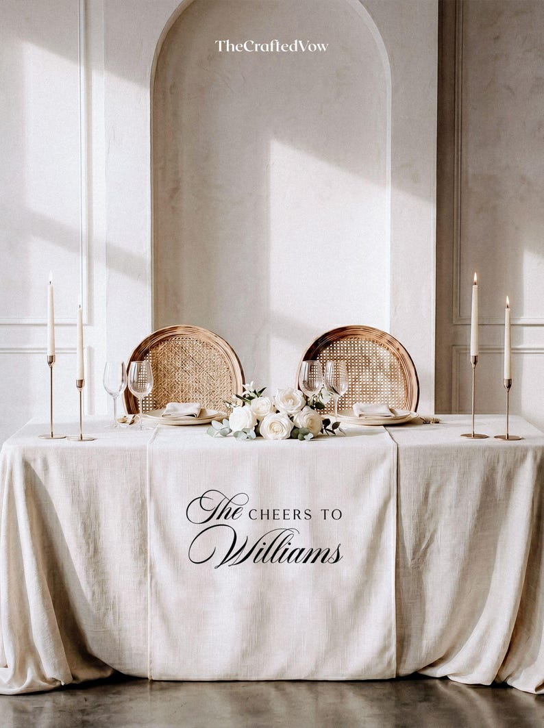 May include: A wedding table setting with a cream tablecloth. Two rattan chairs are placed opposite each other. The table is decorated with white roses, candles, and wine glasses. The text "The Cheers to Williams" is printed on the tablecloth.