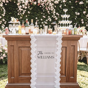 May include: A wooden bar with a white lace runner reading "The Cheers to Williams." The bar is stocked with bottles, glasses, and drinks. A champagne tower is on the right, with a floral backdrop of roses.