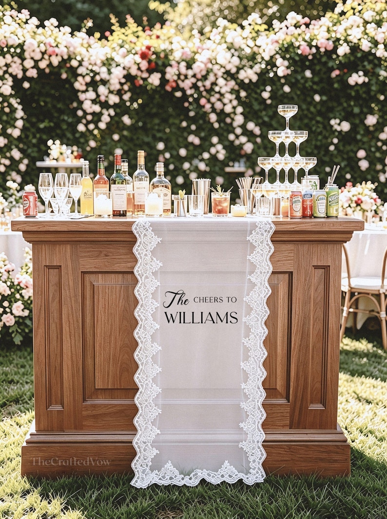May include: A wooden bar with a white lace runner reading "The Cheers to Williams." The bar is stocked with bottles, glasses, and drinks. A champagne tower is on the right, with a floral backdrop of roses.