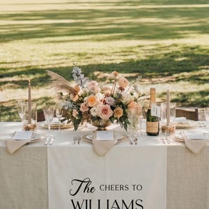 May include: Wedding reception table setting with floral arrangement, champagne, and candles. The table is set with plates, cutlery, and napkins. The table runner reads "The Cheers to WILLIAMS".
