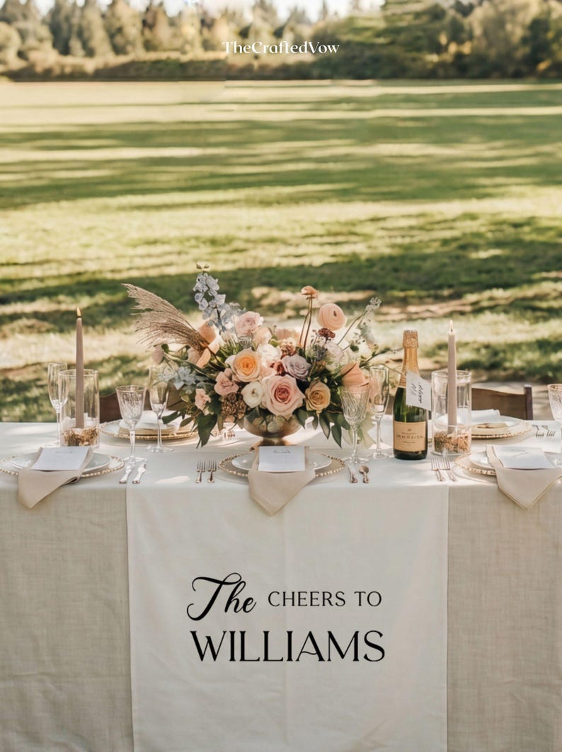 May include: Wedding reception table setting with floral arrangement, champagne, and candles. The table is set with plates, cutlery, and napkins. The table runner reads "The Cheers to WILLIAMS".
