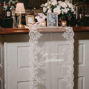 May include: A wedding bar display featuring a white lace runner with intricate floral edging. The text "The Cheers to Williams" is displayed on the runner. The bar is decorated with flowers, candles, framed photos, and a small lamp.