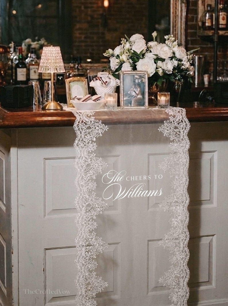 May include: A wedding bar display featuring a white lace runner with intricate floral edging. The text "The Cheers to Williams" is displayed on the runner. The bar is decorated with flowers, candles, framed photos, and a small lamp.