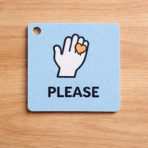 May include: A light blue square coaster featuring a white hand illustration with an orange heart, and the word "PLEASE" in black. The coaster has a small hole at the top and is set on a wooden surface.