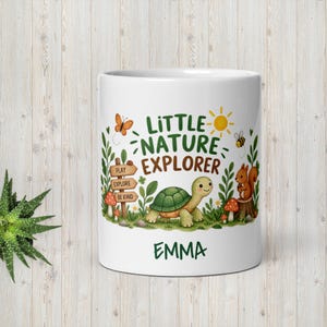 Könnte beinhalten: Ein weißer Keramikbecher mit einer farbenfrohen Illustration einer Schildkröte, eines Eichhörnchens, eines Schmetterlings und einer Biene. Der Text lautet Little Nature Explorer und der Name Emma. Ein Holzschild zeigt Play, Explore, Be Kind.