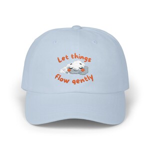 Puede incluir: Gorra de béisbol azul claro con la frase bordada "Let things flow gently" en naranja. La gorra presenta un diseño de nube de dibujos animados con los ojos cerrados y mejillas rosadas. Un accesorio casual.
