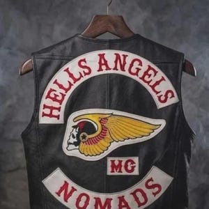 Könnte beinhalten: Eine schwarze Lederweste mit gestickten Aufnähern auf der Rückseite. Der obere Aufnäher zeigt den Schriftzug HELLS ANGELS in roter Schrift auf weißem Grund. Ein zentraler Aufnäher zeigt ein geflügeltes Totenkopf-Logo in Gelb, Rot und Weiß sowie ein MC-Patch.