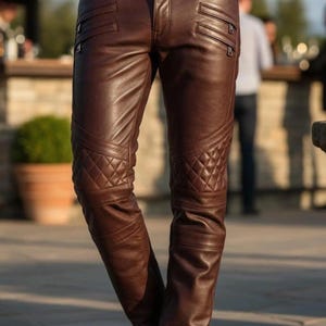 Könnte beinhalten: Braune Lederhose mit gesteppten Knieeinsätzen und Reißverschlusstaschen. Die Hose hat ein klassisches Fünf-Taschen-Design und eine maßgeschneiderte Passform. Das Leder hat ein sattes, glänzendes Finish.