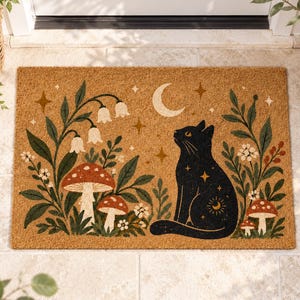 May include: A rectangular tan coir doormat featuring a black cat sitting among green foliage, red and white mushrooms, and white bell-shaped flowers. The design includes a crescent moon and gold-coloured stars scattered across the surface.