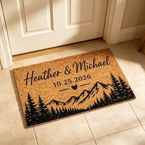 以下が含まれることがあります： 天然ココナッツ繊維で作られた長方形のドアマット。「Heather & Michael」という名前と「10.25.2026」の日付、ハートのシンボル、黒い山と木の模様が特徴です。