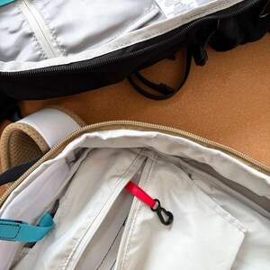 May include: Close-up of two open backpacks. One is black with a gray interior and a teal strap. The other is beige with a white interior and a red zipper pull. Both have zippers and black hardware.