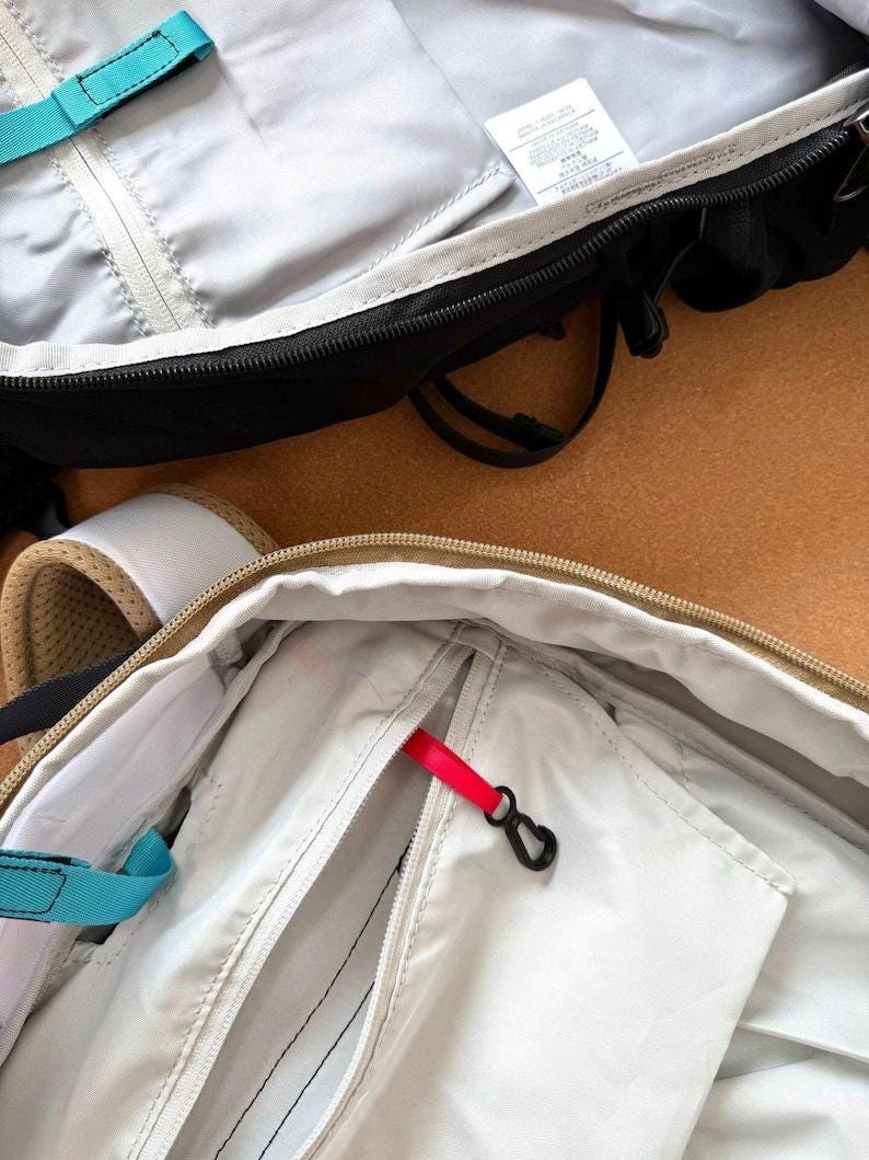 May include: Close-up of two open backpacks. One is black with a gray interior and a teal strap. The other is beige with a white interior and a red zipper pull. Both have zippers and black hardware.