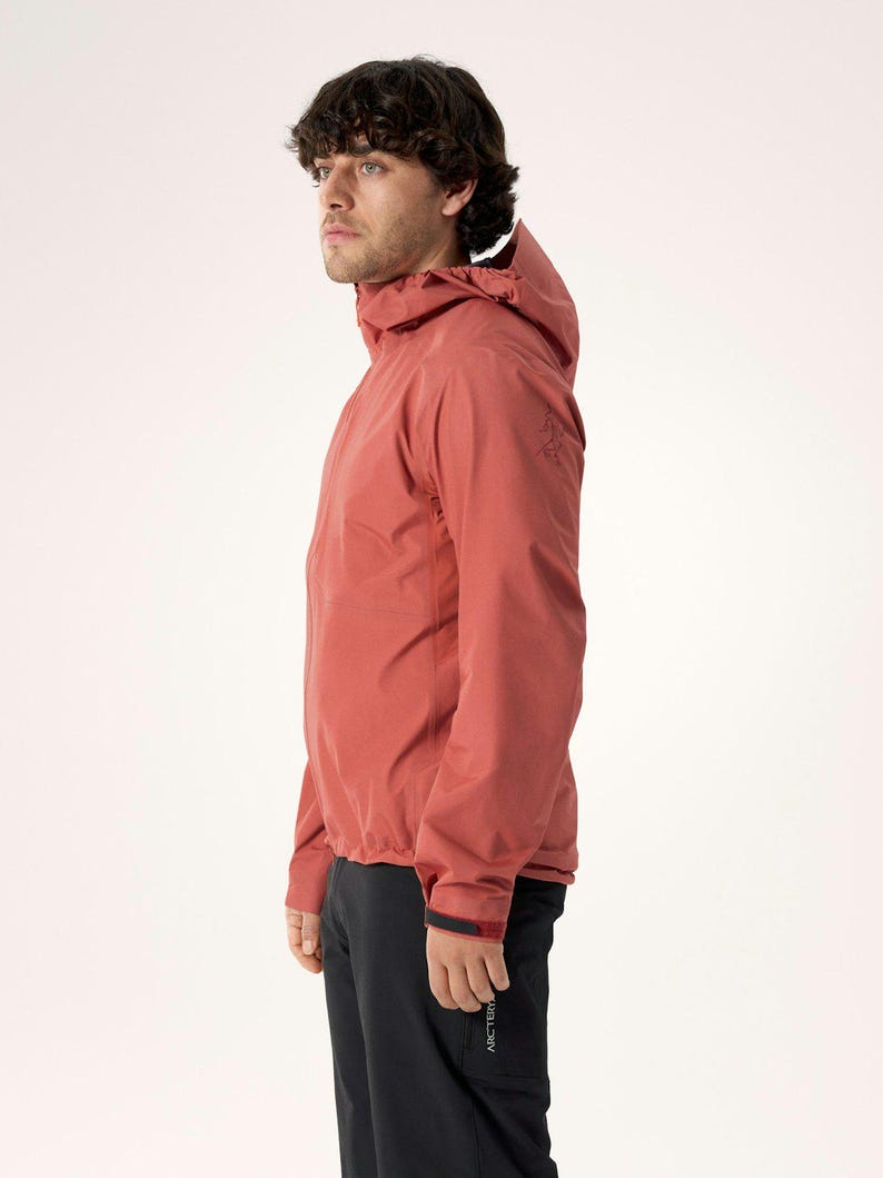 Puede incluir: Chaqueta con capucha de color &oacute;xido con cierre de cremallera. La chaqueta tiene un dise&ntilde;o estilizado y se combina con pantalones negros. El modelo est&aacute; mirando hacia la izquierda, mostrando el perfil lateral de la chaqueta. La chaqueta parece estar hecha de un material ligero y resistente al agua.