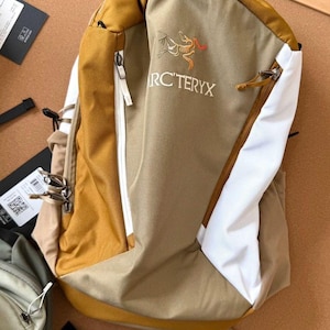May include: A tan and white backpack with a mustard yellow top and accents. The backpack has a zippered front pocket and the text "ARC'TERYX" is visible. The backpack is made of a durable material.