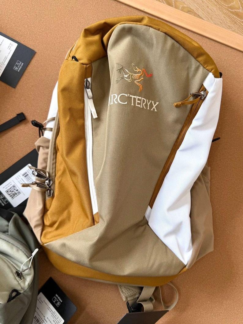 May include: A tan and white backpack with a mustard yellow top and accents. The backpack has a zippered front pocket and the text "ARC'TERYX" is visible. The backpack is made of a durable material.