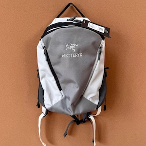 May include: A gray, white, and black backpack with the Arc'teryx logo. The backpack has a top handle, two side pockets, and adjustable straps. The bag is hanging against a brown background.