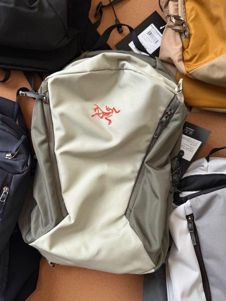 May include: A light beige and gray backpack with a red logo on the front. The backpack has two zippered compartments and a drawstring closure. Several other backpacks are visible in the background.
