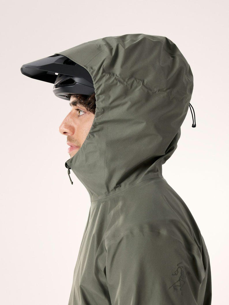Puede incluir: Una persona con una chaqueta con capucha verde oscuro y un casco de bicicleta negro. La chaqueta tiene un cuello alto y una capucha con cord&oacute;n. Un peque&ntilde;o logotipo es visible en la manga izquierda.
