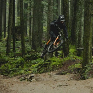 Puede incluir: Un ciclista de monta&ntilde;a con equipo negro salta con su bicicleta de monta&ntilde;a naranja y negra en un bosque. La escena est&aacute; rodeada de altos &aacute;rboles verdes y suelo cubierto de musgo, creando una imagen din&aacute;mica de aventura al aire libre.