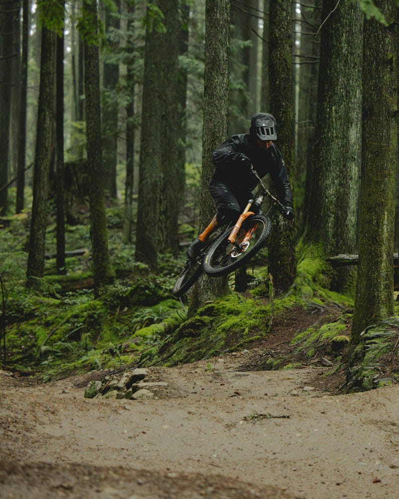 Puede incluir: Un ciclista de monta&ntilde;a con equipo negro salta con su bicicleta de monta&ntilde;a naranja y negra en un bosque. La escena est&aacute; rodeada de altos &aacute;rboles verdes y suelo cubierto de musgo, creando una imagen din&aacute;mica de aventura al aire libre.