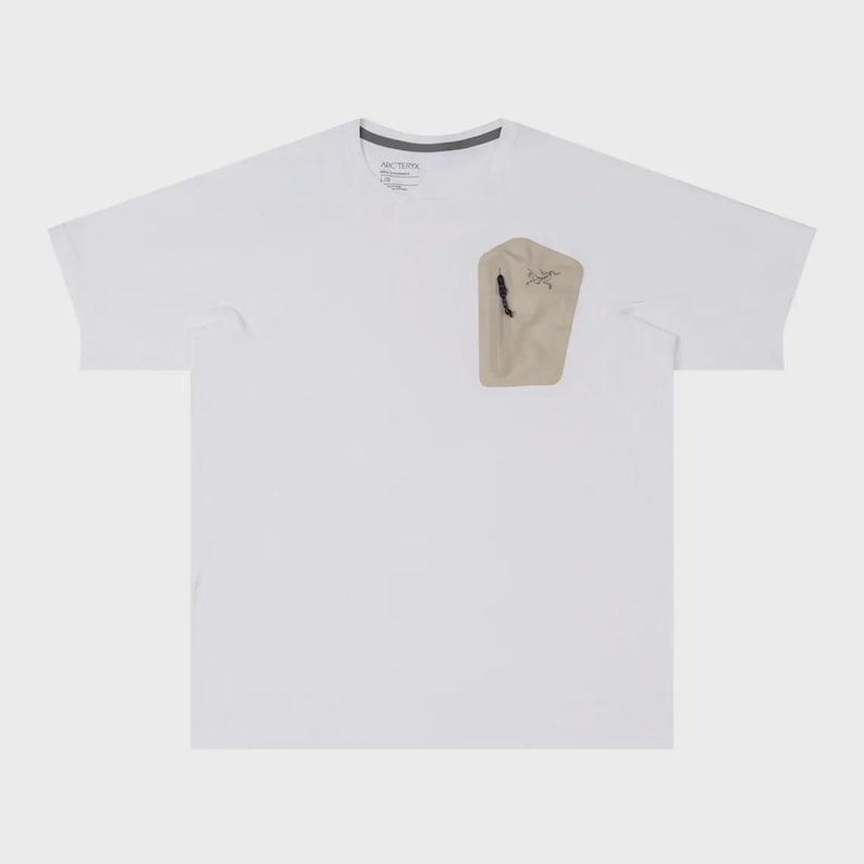 May include: White short-sleeve t-shirt with a beige pocket on the chest. The pocket has a black zipper and a small logo. The shirt has a crew neck and a tag at the neckline.