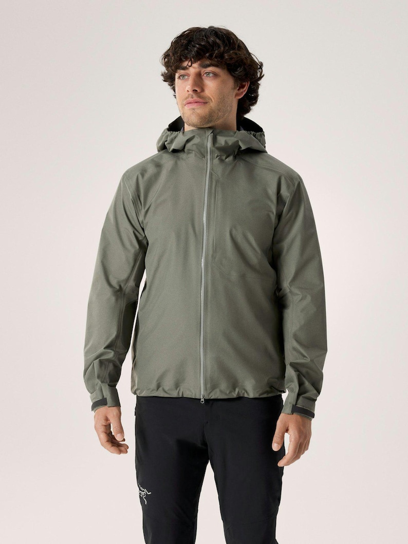 Puede incluir: Chaqueta con capucha verde salvia con cremallera completa, usada con pantalones negros. La chaqueta tiene un dise&ntilde;o aerodin&aacute;mico con capucha y pu&ntilde;os. El modelo est&aacute; de pie frente a un fondo liso.