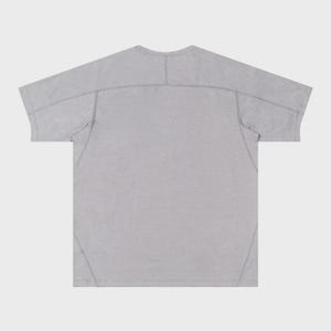 May include: A light gray t-shirt with a crew neck and short sleeves. The back of the shirt is visible, showing seams across the shoulders and down the sides. The fabric has a subtle texture.