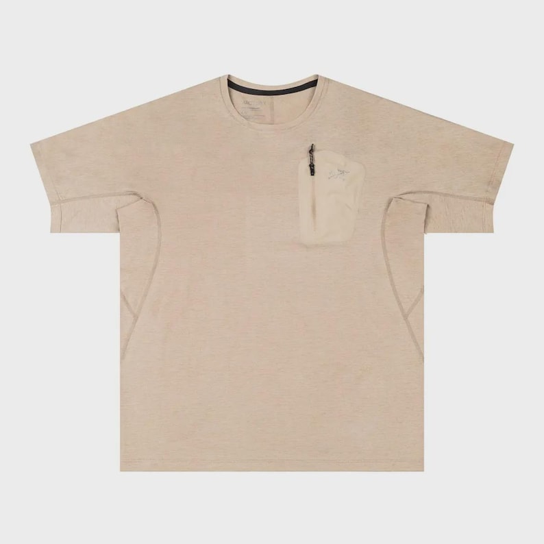 May include: A beige short-sleeved t-shirt with a small pocket on the chest. The pocket has a zipper and a small design. The shirt has a crew neck with a black trim. The fabric appears to be lightweight.