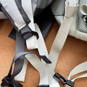 May include: Close-up of a backpack with gray and white straps and black plastic buckles. The straps are made of a durable, woven material. The backpack appears to be new and unused.