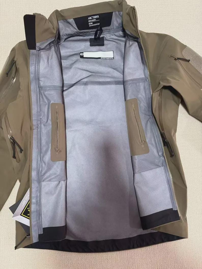 Może przedstawiać: Otwarta kurtka outdoor w kolorze piaskowym z szarą podszewką. Kurtka posiada liczne kieszenie zapinane na zamek, kaptur oraz nazwę marki "Arc'teryx" na kołnierzu. Zaprojektowana do aktywności na świeżym powietrzu.