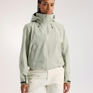 Puede incluir: Chaqueta con capucha verde claro con cremallera completa, combinada con pantalones color crema. La chaqueta tiene un cuello alto y detalles negros en los puños. La modelo está frente a un fondo blanco. La chaqueta parece estar hecha de material impermeable.