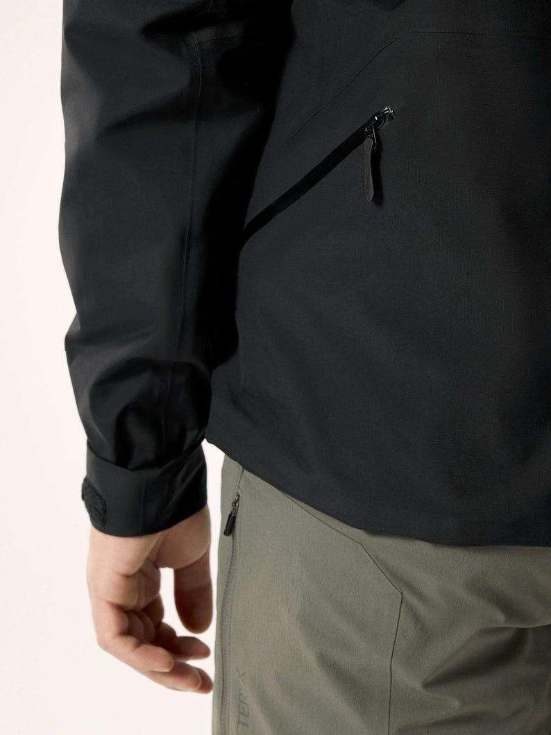 Puede incluir: Chaqueta negra con bolsillo con cremallera y pu&ntilde;o con cierre de velcro. La chaqueta se combina con pantalones grises con bolsillo con cremallera. La imagen muestra un primer plano de la chaqueta y los pantalones.