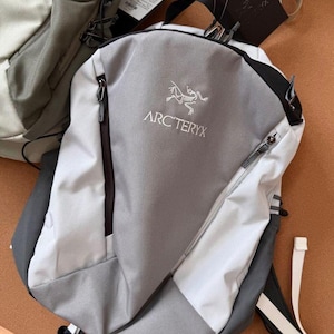 May include: A gray and white backpack with the Arc'teryx logo. The backpack has a zippered main compartment and a smaller front pocket. The straps are black and white. The backpack is made of a durable, water-resistant material.