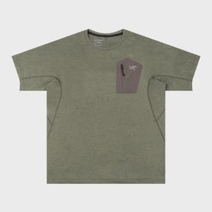 May include: Olive green short-sleeved t-shirt with a small, rectangular pocket on the chest. The pocket is a muted brown color and features a white logo and text. The shirt has a crew neck and a relaxed fit.