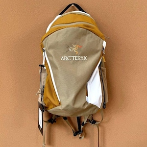 May include: A tan and mustard-colored backpack with the Arc'teryx logo. The backpack has white accents, a black handle, and adjustable straps. It features multiple compartments and a zipper closure. A tag hangs from the bottom.