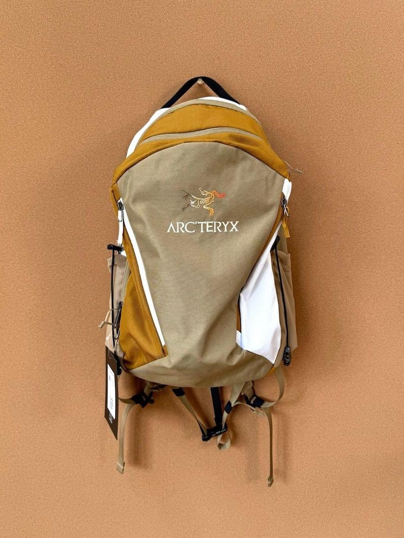 May include: A tan and mustard-colored backpack with the Arc'teryx logo. The backpack has white accents, a black handle, and adjustable straps. It features multiple compartments and a zipper closure. A tag hangs from the bottom.