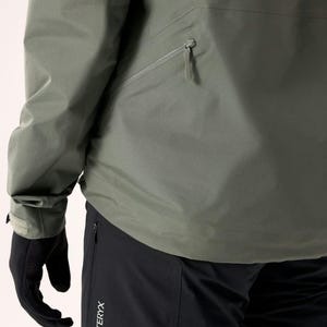 Puede incluir: Primer plano de una chaqueta verde salvia con un bolsillo en el pecho con cremallera y un pu&ntilde;o en la manga. La chaqueta se combina con pantalones negros. El logotipo de Arc'teryx es visible en los pantalones. La persona lleva guantes negros.