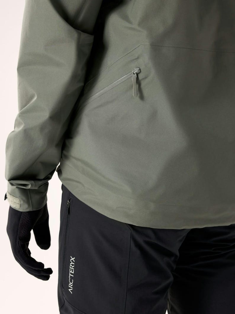 Puede incluir: Primer plano de una chaqueta verde salvia con un bolsillo en el pecho con cremallera y un pu&ntilde;o en la manga. La chaqueta se combina con pantalones negros. El logotipo de Arc'teryx es visible en los pantalones. La persona lleva guantes negros.