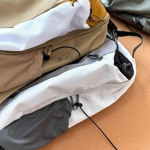 May include: A close-up of several backpacks in various colors, including tan, white, and gray. The backpacks have multiple compartments, zippers, and straps. The word "ARCTERYX" is visible on one backpack, and "BEAMS" is visible on another.