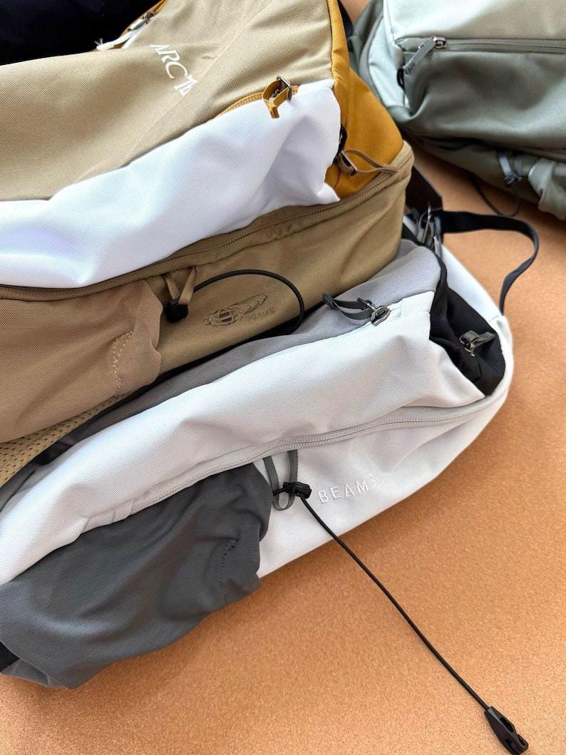 May include: A close-up of several backpacks in various colors, including tan, white, and gray. The backpacks have multiple compartments, zippers, and straps. The word "ARCTERYX" is visible on one backpack, and "BEAMS" is visible on another.