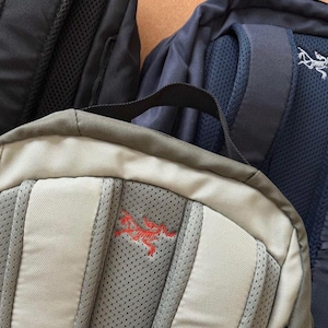 May include: Close-up of three backpacks in various colors. The front-most backpack is tan with a red logo. The other two backpacks are black and navy blue. All three backpacks have padded backs and shoulder straps.