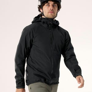 Puede incluir: Una chaqueta negra con capucha y cremallera completa, combinada con pantalones verde oliva. La persona lleva un casco negro. La chaqueta parece estar hecha de un material ligero e impermeable, adecuado para actividades al aire libre.