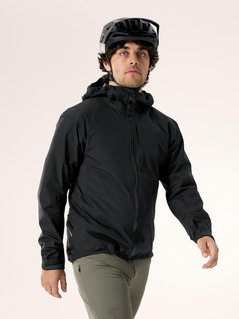 Puede incluir: Una chaqueta negra con capucha y cremallera completa, combinada con pantalones verde oliva. La persona lleva un casco negro. La chaqueta parece estar hecha de un material ligero e impermeable, adecuado para actividades al aire libre.