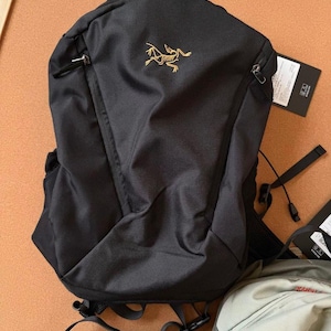 May include: Black backpack with a gold logo on the front. The backpack has a top handle, two zippered pockets, and adjustable straps. A tag is attached to the side. The backpack is made of a durable, water-resistant material.