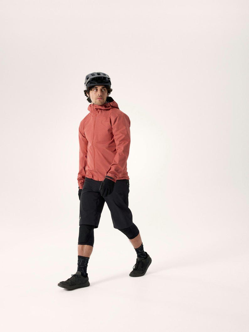 Puede incluir: Una persona con una chaqueta con capucha de color coral, pantalones cortos negros, guantes y zapatos, con un casco negro. El atuendo incluye rodilleras negras y calcetines estampados. La persona camina sobre un fondo blanco.